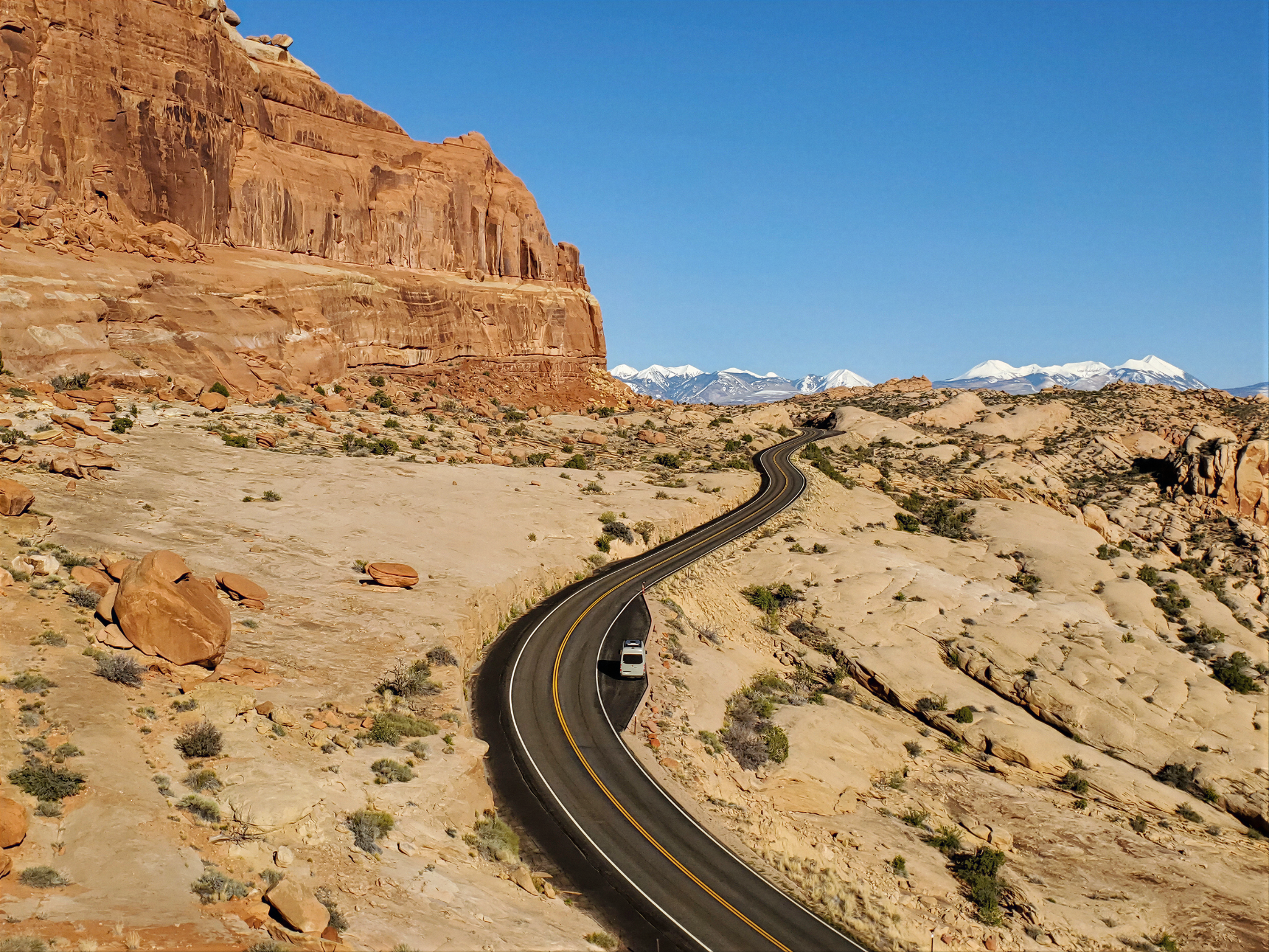 A Nokhu camper van traveling along a winding desert road between sandstone cliffs and distant snow-capped mountains